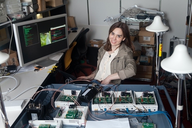 Photo of a woman sitting before a computer monitor, next to a tray full of electronic components and cables
