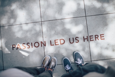 Two pairs of sneakered feet on the sidewalk with the words PASSION LED US HERE