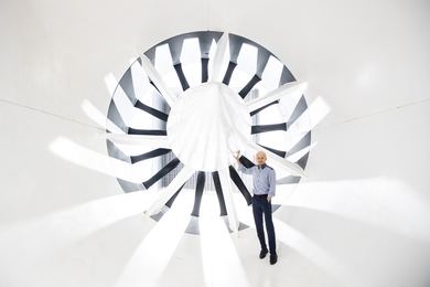 Photo of Mark Drela standing before a large fan inside a white wind tunnel. The scene is backlit with rays of light radiating dramatically toward the camera.