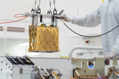 The MOXIE experiment, in a gold-colored housing about a foot cubed, is handled by a person wearing a white clean suit in a clean room
