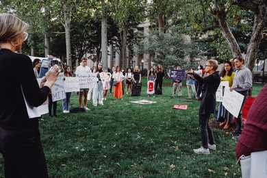 Photo of Linda Griffith with a bullhorn, addressing a semicircle of about 30 rally attendees on a grassy area, many holding hand-painted signs.