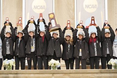 Nine women in identical caps and warmup jackets pose in a row, hoisting trophies over their heads