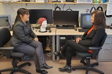 Diane Chan and Li-Huei Tsai sit, talking, at a table covered in research equipment. In front of powered-off monitors sits a polystyrene head with a red cap of electrodes