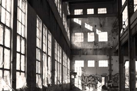 Nighttime black and white photo outside a warehouse where the lights are on, and the exterior wall has been marked with graffiti 