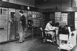 Black-and-white photo showing Jack Gilmore standing next to the Whirlwind computer, while Joe Thompson sits, working on the machinery