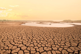 Photo of a dried-up lakebed with cracked mud taking up most of the scene, with a small pond of water in the distance