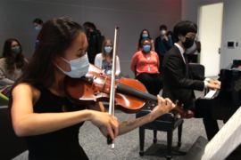 Photo of a woman playing violin and a man playing piano in a small room. About half a dozen people are seated behind them. Everyone is wearing face masks.