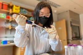 Photo of Anna Yeh wearing goggles, a lab coat, a mask, and gloves and uses a pipette in a lab