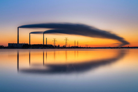 three smokestacks billow symmetric plumes of smoke over the horizon, which is reflected in a pool of still water below.