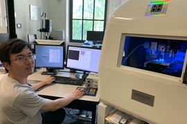 Photo of Jiahao Dong sitting at a desk with two monitors in Jaramillo lab. A large piece of lab equipment labeled “TriboIndenter” is in the foreground.