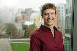 Amy Keating stands in a red button-down blazer in front of her office window at MIT.
