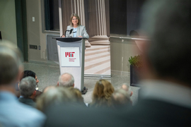 Sally Kornbluth speaks at a podium in front of a blackboard, while audience members watching are seen in the foreground
