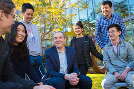 David Autor, seated outside in front of a tree and a building with glass windows, is surrounded by six young adults, all of whom are smiling.