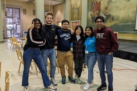 Six students stand with their arms around one another, one giving a thumbs-up