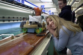Woman leans on the ledge surrounding an indoor water tank, posing next to a small robotic boat that is also resting on the ledge
