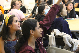 About a dozen students seated at tables in side-view. One student has a raised hand.