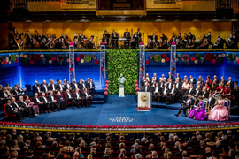 Overview of a large stage with many Nobel Prize winners and guests, all in formal dress, plus the king and queen of Sweden and an orchestra above.