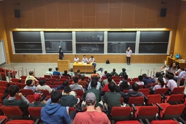 A semi-full lecture hall, photographed from the back of the room. Two teams of four high school students sit in the first row. Facing the audience in front of a large set of chalkboards are five volunteers in purple keeping score, operating the timer, and reading questions.