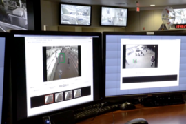 In the foreground, two desktop monitors display surveillance videos of a subway and a street scene. Two more monitors are mounted near the ceiling in the background