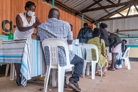 Several people, seen from the back, sit in plastic chairs at a table while health care workers inject vaccine.