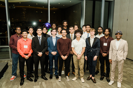 About 20 young men of color pose for a photo in front of a wall of windows at night
