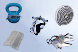 Five objects appear on a whitish background: A blue kettlebell with some microchip components, a silver ringlike object with wires, a roll of corrugated white plastic, a white glove with wires on it, and an off-white corrugated piece of plastic. 