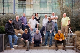 12 people pose on outdoor steps in front of a wall of windows, some sitting and others standing