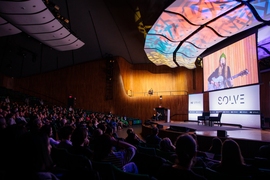 A woman seated on a well-lit balcony plays guitar and sings to a crowd in a dimly lit auditorium. Her image is projected on a screen over the main stage.