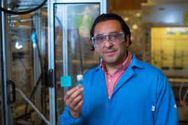 Yuriy Román-Leshkov, wearing safety glasses and a lab coat, holds vial of clear liquid up for the camera. 
