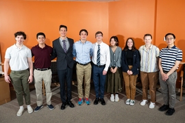 Nine interns pose for a group photo in front of an orange wall. 