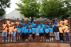 About 50 dynaMIT and MIT students wearing matching blue or orange T-shirts pose in three rows on the steps of the Stata Center
