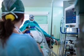 A hospital operating room is seen from behind a blue-gowned anesthesiologist as she applies a mask to a patient. Another doctor is at the far end of the gurney.