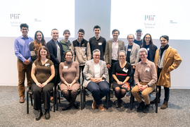 16 people pose in two rows, one seated and one standing, for a group photo in MIT's Hayden Library.
