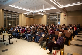 In a small auditorium, about 100 people sit listening to a speaker who is out of frame