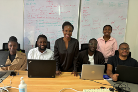 Six people pose behind a table. Two are standing, and the seated ones each have a laptop