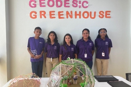 Five children wearing purple shirts stand against a wall displaying the words “Geodesic Greenhouse.” Two geodesic models are on a table in front of them.
