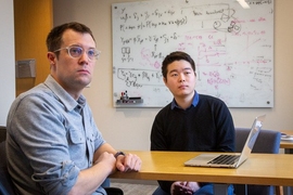 Two men sitting at a table, with a whiteboard in the background. 