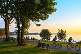 About two dozen people walking, biking, and relaxing in a verdant park next to a lake on a sunny summer day