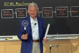 Male instructor teaches in front of a blackboard that contains a series of terms including "plasma ion source," and "collision reaction cell."  He is holding a small round lightbulb in one hand and a long fluorescent lightbulb in the other.