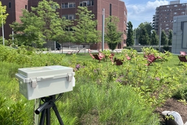 a tripod with small gray plastic box on top overlooks a lawn with chairs and buildings in the background