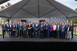 36 individuals pose on an outdoor stage under a pyramidal awning