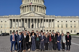 26 people pose together in front of the U.S. Capitol on a sunny day.