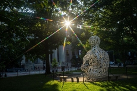 Image of The Alchemist sculpture on MIT campus with trees and sun shining through.