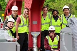 Seven men wearing safety yellow work vests and hardhats pose by a metal pillar that has a small gray patch in its center.
