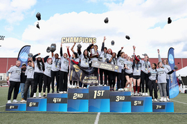 About 20 MIT women's team members and three coaches celebrate on the podium by throwing their hats in the air 
