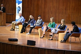 Six panelists seated on a stage, with a moderator visible at a lectern to their right