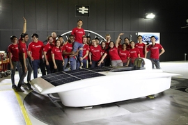 22 people in matching red T-shirts pose behind a solar electric vehicle. In the middle of the line, two men boost up Francis Wang