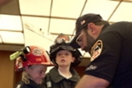 World Trade Center Task Force Fire Safety Director Mike Bellone helps Jamie Joyce, 8, into one of the jackets he and his fellow workers wore in their rescue efforts at the World Trade Center. Jamie's brother Patrick, 10, stands nearby, outfitted in a similar jacket and hat. The children's great-grandfather was a Somerville firefighter. Their great-aunt Kathy Doyle works at Sloan School.