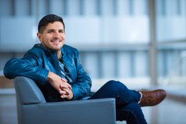 Vila sits on a blue chair with legs crossed, and large windows in background.