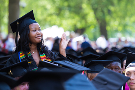 A female graduate standing above the black mortarboard hats of her seated fellow graduates.